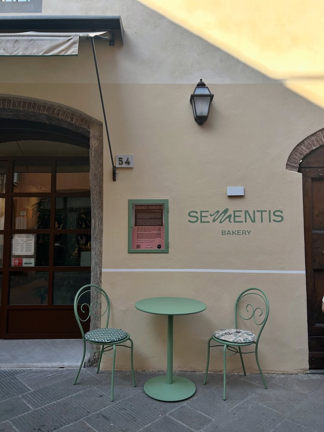 The sage-green storefront of Sementis Bakery in Pietrasanta, with a small table and two chairs outside.